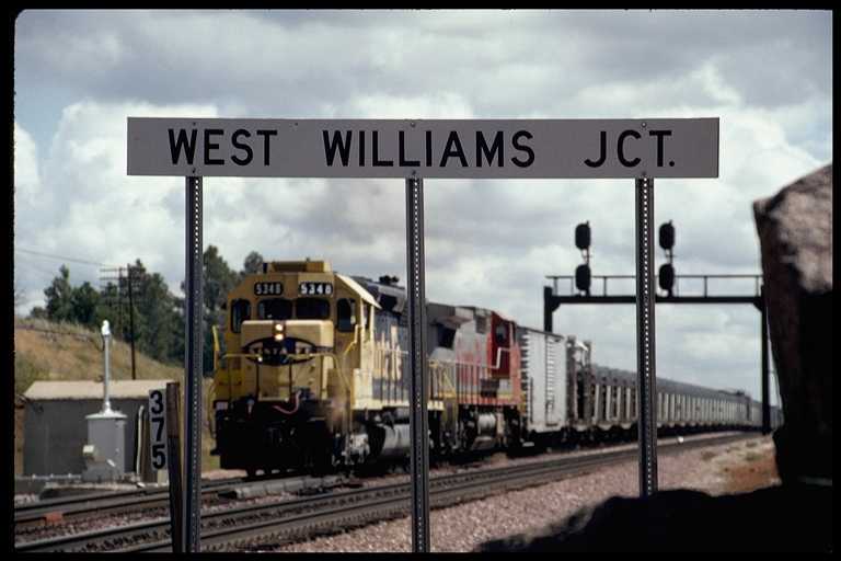 West bound train passing the West Williams Jct. sign. Train is carrying rail.