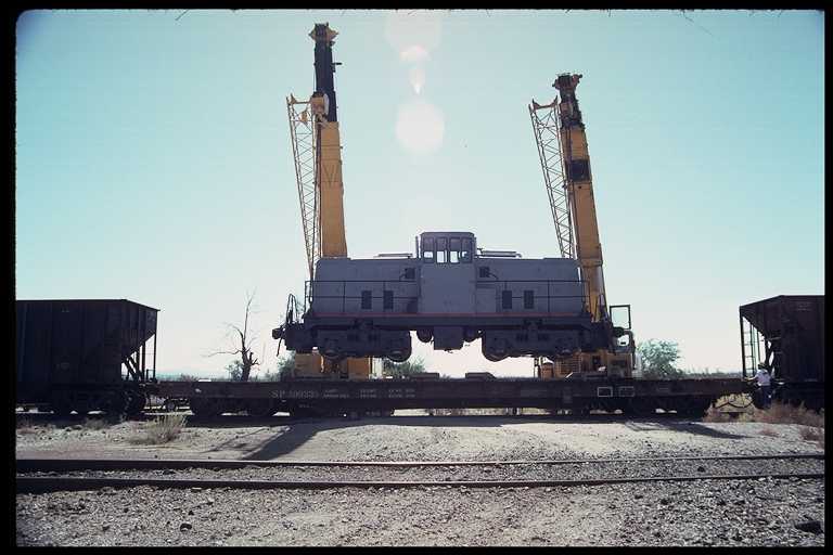 Center cab locomotive being delivered by flatcar.  Locomotive being lifted by twp cranes.